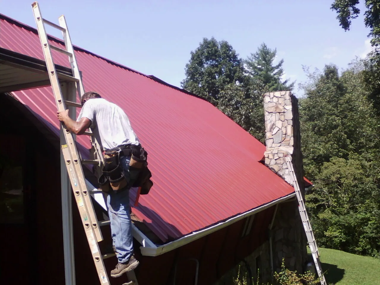 Roofer on a ladder beside a red metal roof and stone chimney — Soffit & Fascia Repair by Roofing Brothers in Sumas